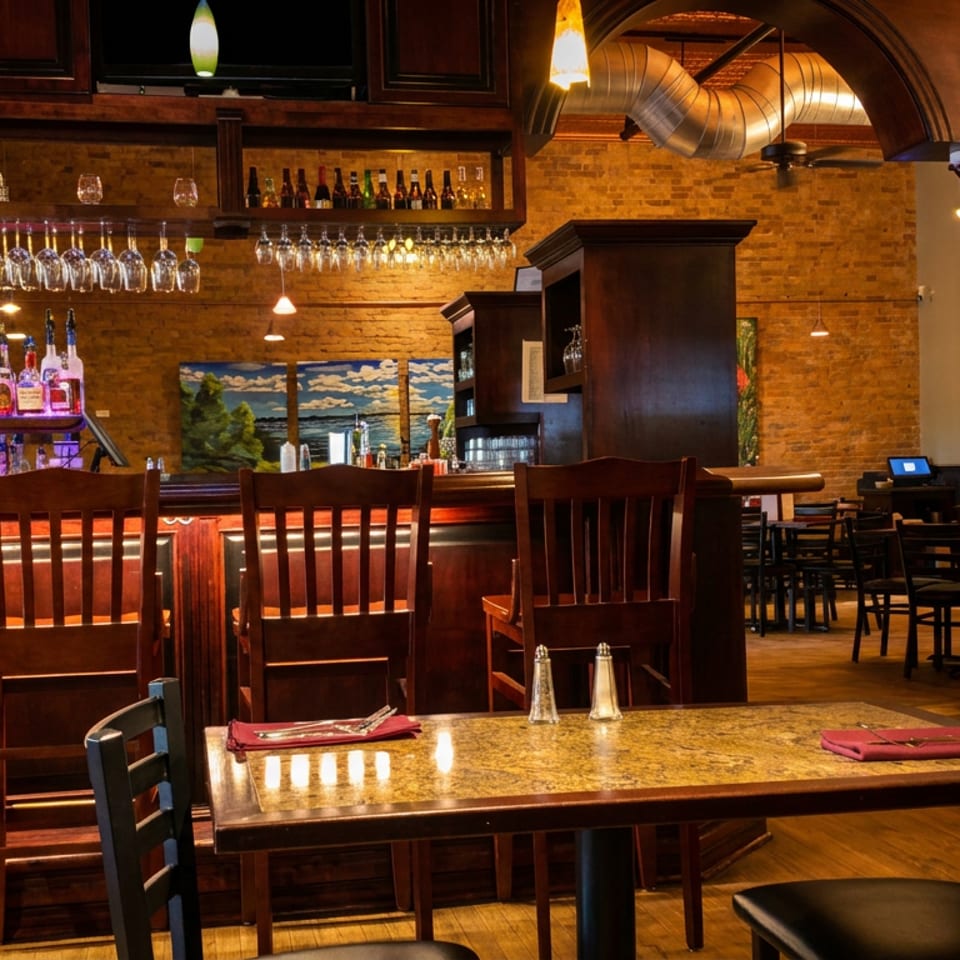 Warmly lit restaurant interior with a full bar, high-top chairs, and dining tables with brick walls.