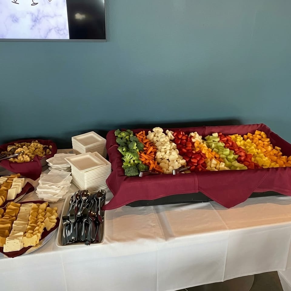 Buffet table featuring colorful cut vegetables, broccoli, cauliflower, carrots, cheese, crackers, plates, napkins, and serving utensils.