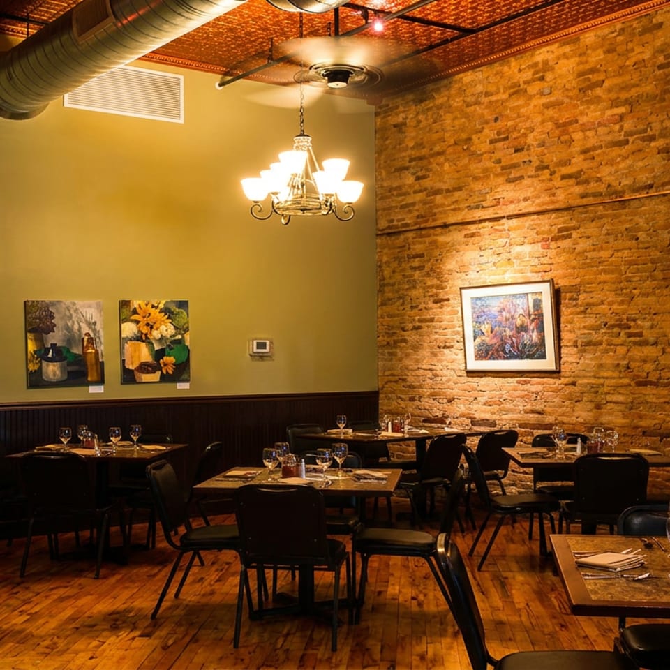 Restaurant dining room with set tables, hardwood floors, exposed brick, and olive green walls with artwork.