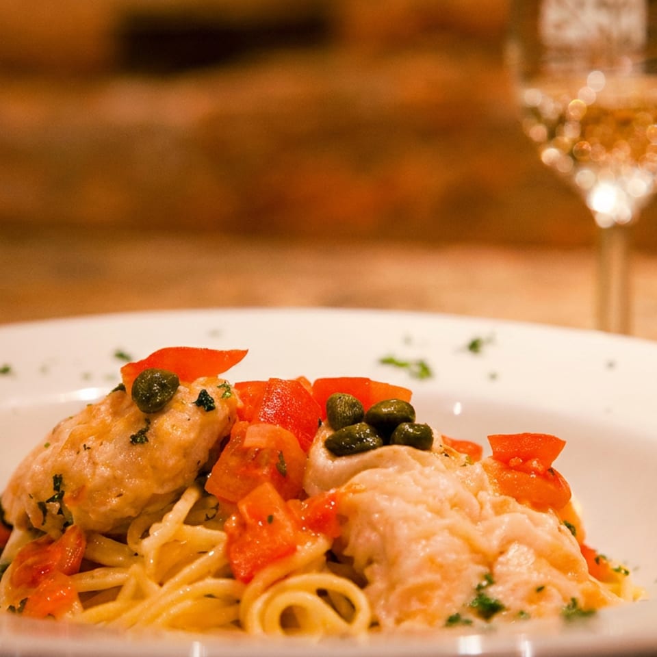 Spaghetti with chicken, diced tomatoes, and capers on a white plate with a glass of wine.