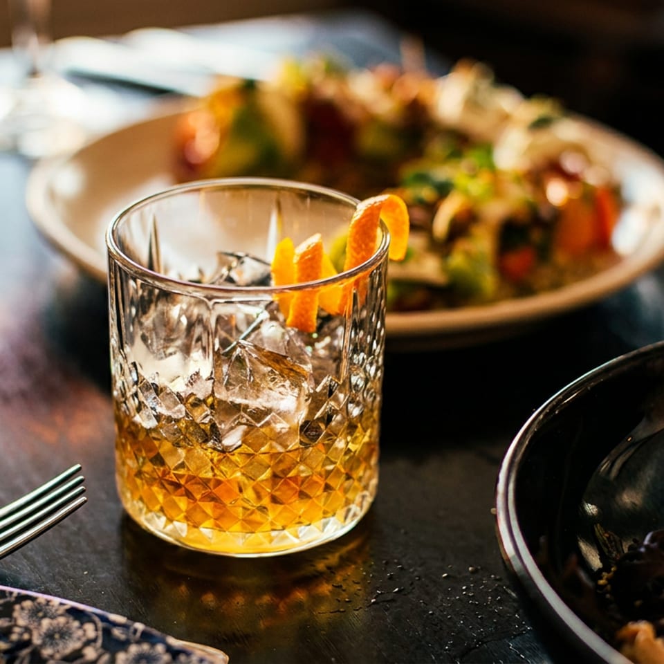 A cocktail with ice and orange peel, with two plates of food on a dark table.