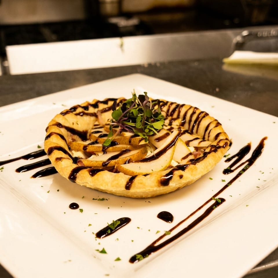 Pear tart with balsamic glaze and microgreens on a white plate in a commercial kitchen.
