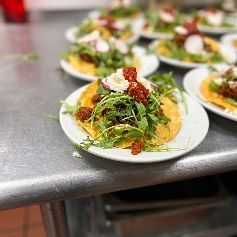 Plated tostadas topped with seasoned filling, arugula, creamy cheese, radishes, and red sauce on a stainless-steel counter.