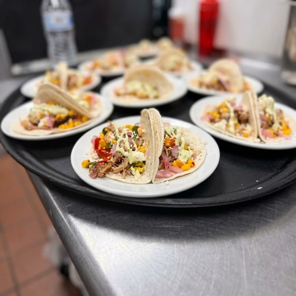 Plates of soft tacos filled with grilled meat, corn, vegetables, pickled onions, and creamy sauce arranged on a serving tray.
